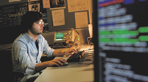 A student on a computer in the background, a computer screen with lines of code in the foreground.