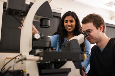 A man and woman looking into a microscope in a lab.                    