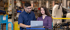 A man and a woman in a factory warehouse working on a laptop computer.