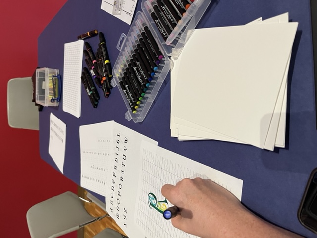 A persons hand writing on a how to sheet of calligrpahy on a table full of markers.