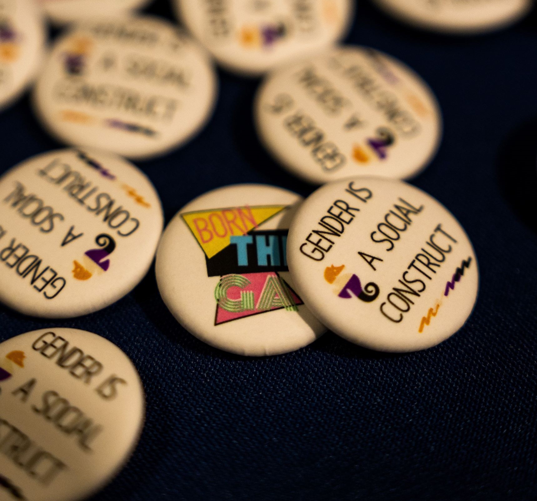 A picture of a pile of pins on a black background. The pins say "GENDER IS A SOCIAL CONSTRUCT" and "BORN THIS GAY"