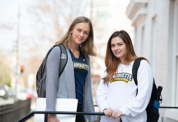 Two women, wearing Marquette branded shirts, standing outside