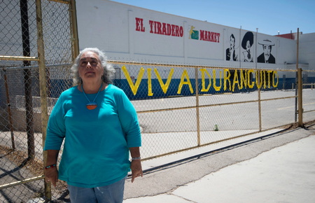 photo of a middle-aged woman standing in front of a low chain-link fence that bears a sign Viva Duranguito. Behind the fence is a building with a painted sign that says El Tiradero Market