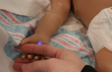 A newborn mannequin grasping a nurse’s finger during