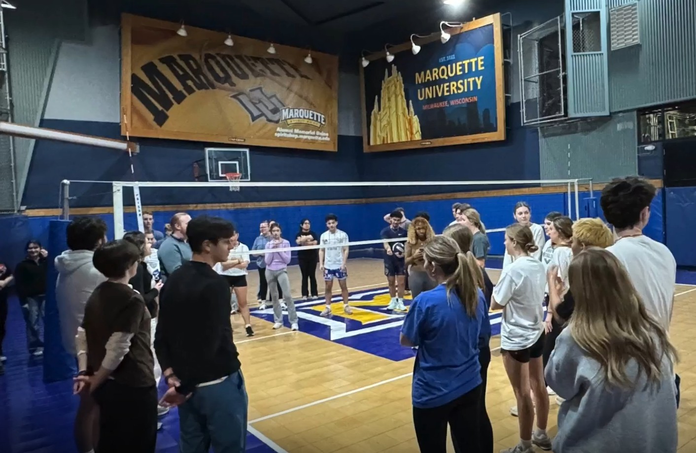group of students gathered around a volleyball net in a gym