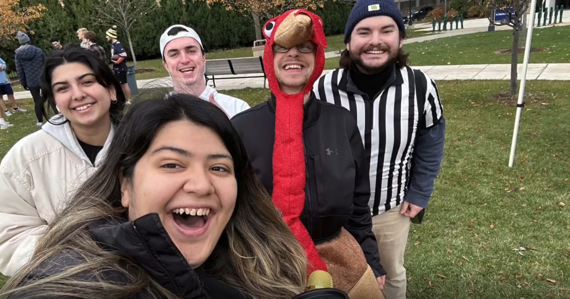 five people smiling at camera while outside in grassy field, one person is wearing a turkey costume, one is in a referee outfit